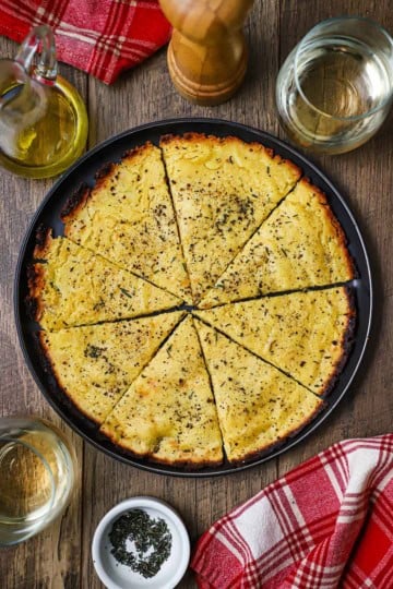 An overhead view of easy homemade socca that has been cut into triangles sitting in a large black plate next to two glasses of white wine and a red checkered napkin.