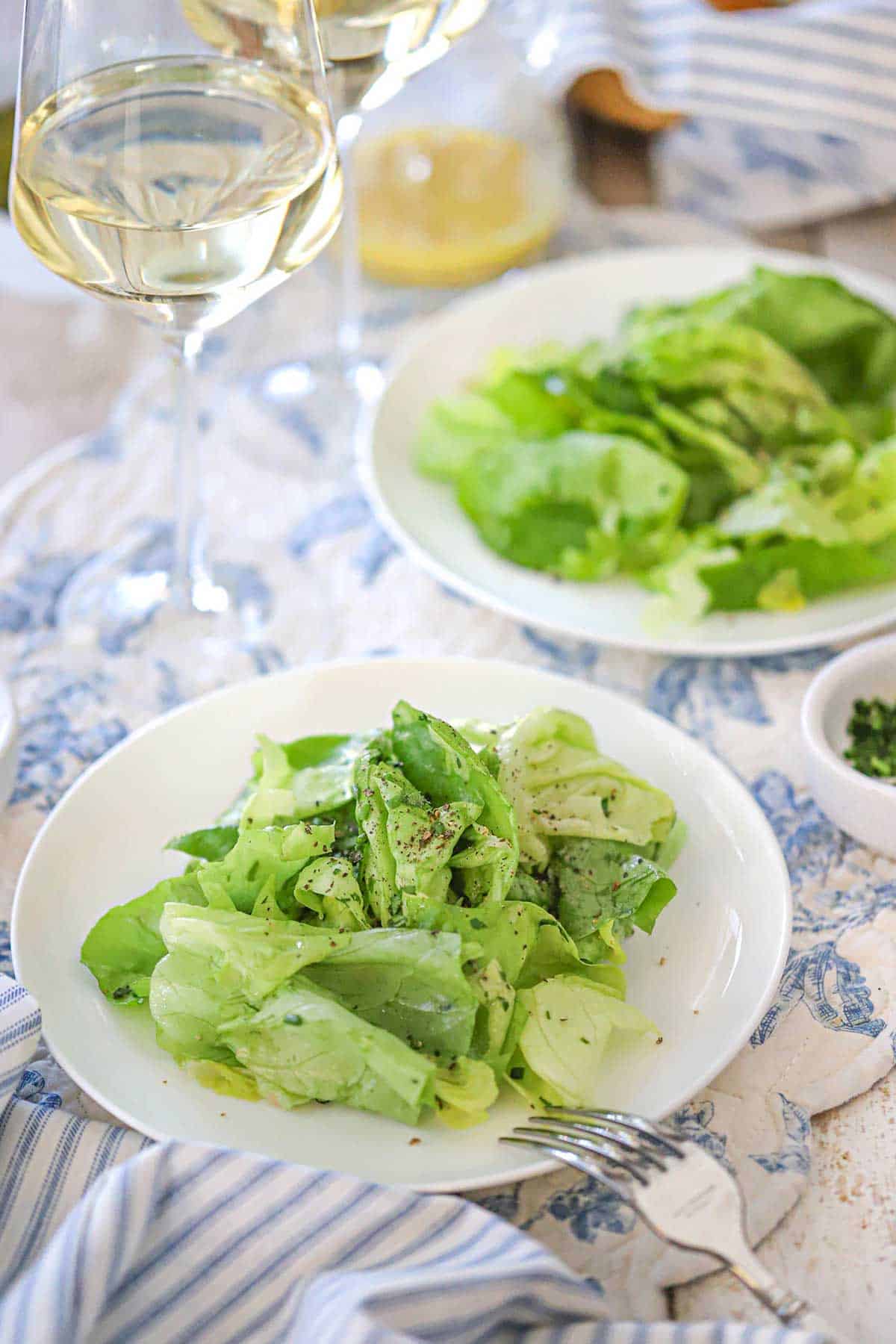 Two white salad plates filled with a serving of Classic French Bistro Salad with two glasses of white wine nearby.