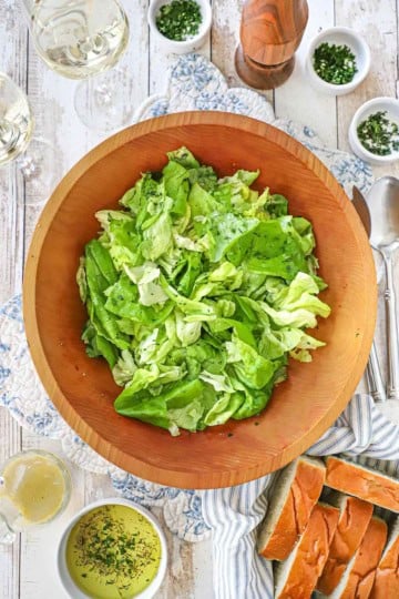 An overhead view of a large wooden salad bowl filled with Classic French Bistro Salad surrounded by small bowls of snipped herbs, sliced French bread, and two glasses of white wine.