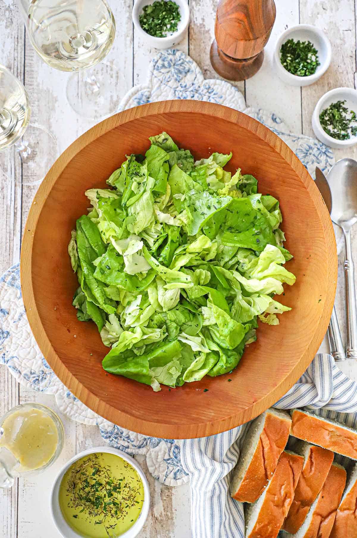 An overhead view of a large wooden salad bowl filled with Classic French Bistro Salad surrounded by small bowls of snipped herbs, sliced French bread, and two glasses of white wine.