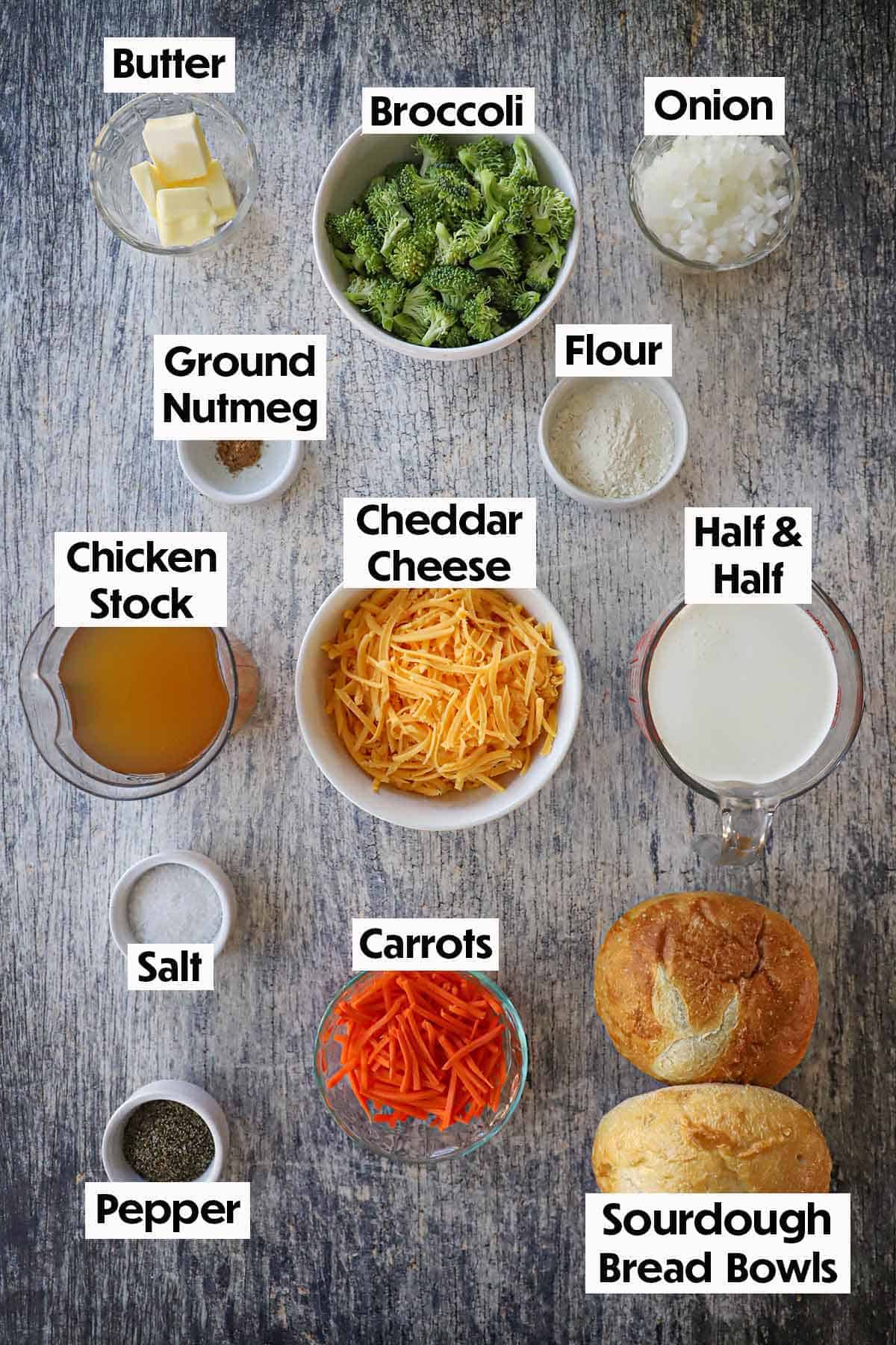An arrangement of ingredients for Broccoli Cheddar Soup (Panera Copycat) on a grey wooden background including shredded cheddar cheese, chopped broccoli, onion, butter, chicken stock, half and half, carrots, bread bowls, and seasonings.