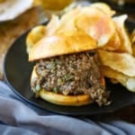 A close-up view of a black dinner plate filled with a stuffed Ultimate Philly Cheesesteak Sloppy Joe next to a pile of homemade potato chips with a glass of beer nearby.
