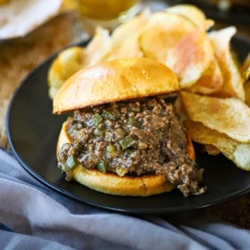 A close-up view of a black dinner plate filled with a stuffed Ultimate Philly Cheesesteak Sloppy Joe next to a pile of homemade potato chips with a glass of beer nearby.