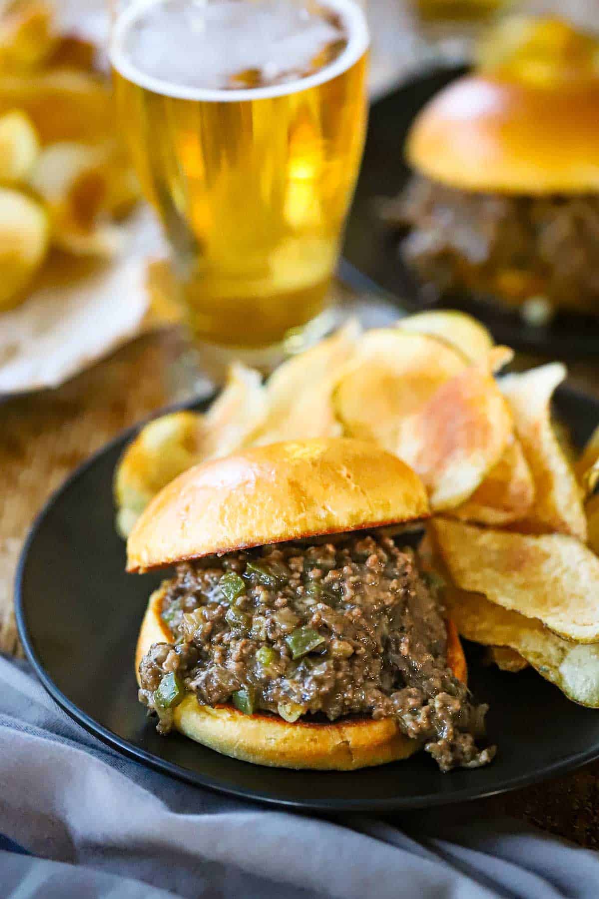 A black dinner plate filled with a stuffed Ultimate Philly Cheesesteak Sloppy Joe next to a pile of homemade potato chips with a glass of beer nearby.