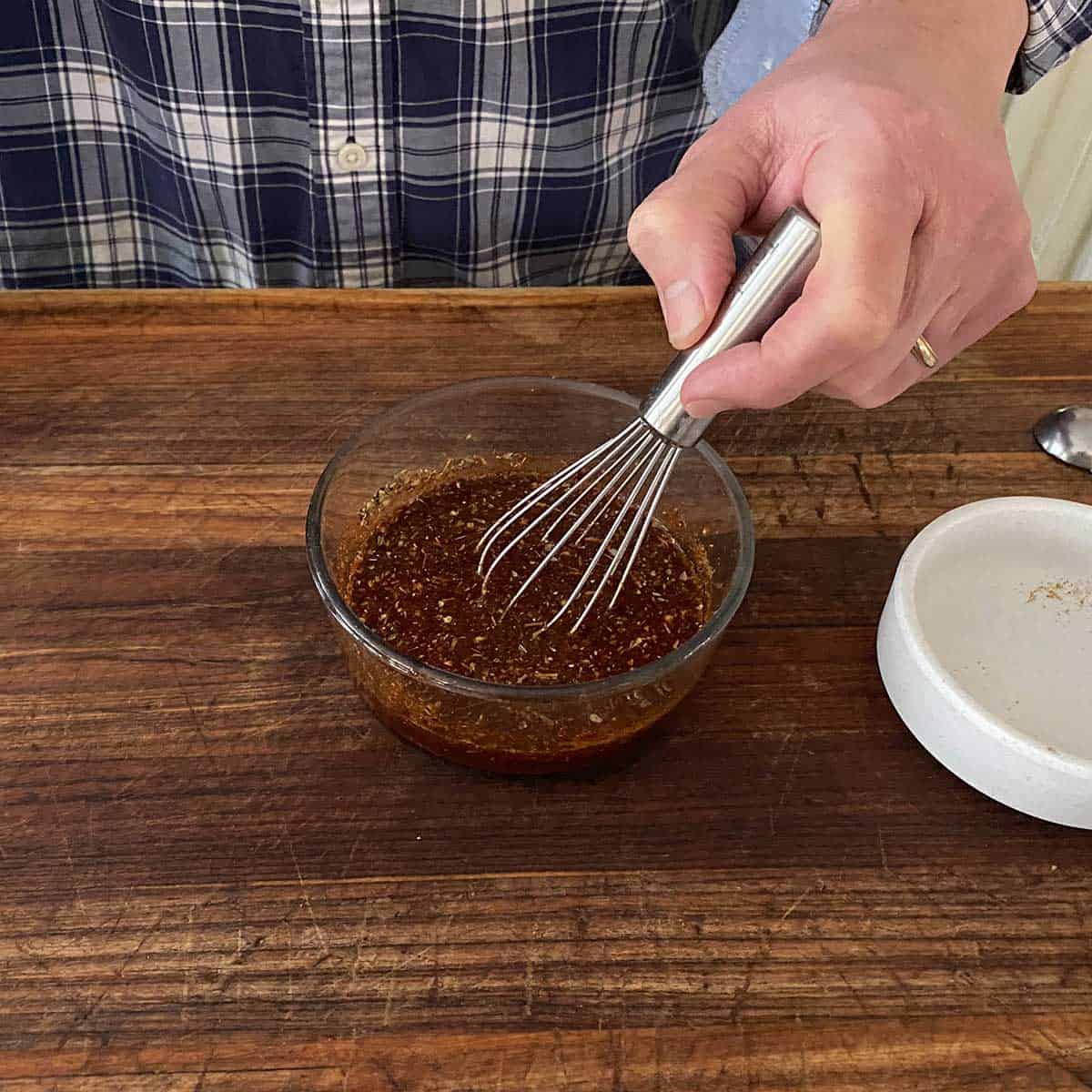 A person using a small whisk to mix a brown marinade for authentic Peruvian chicken on a wooden cutting board.