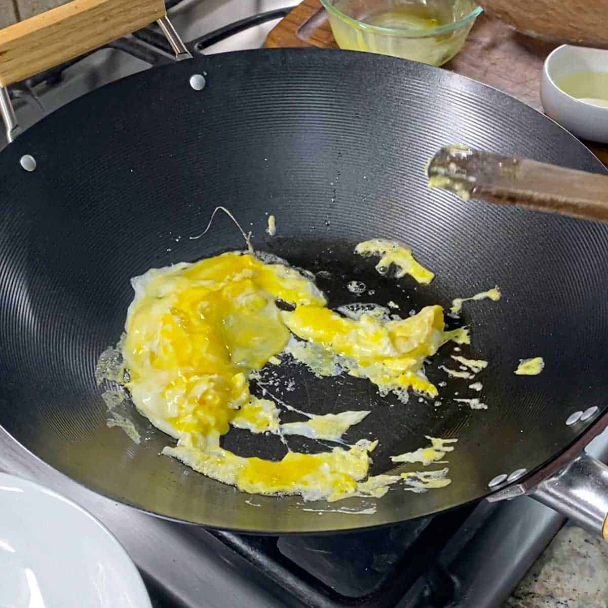 A person using a wooden spatula to stir lightly scrambled eggs in a large wok over a gas stove.