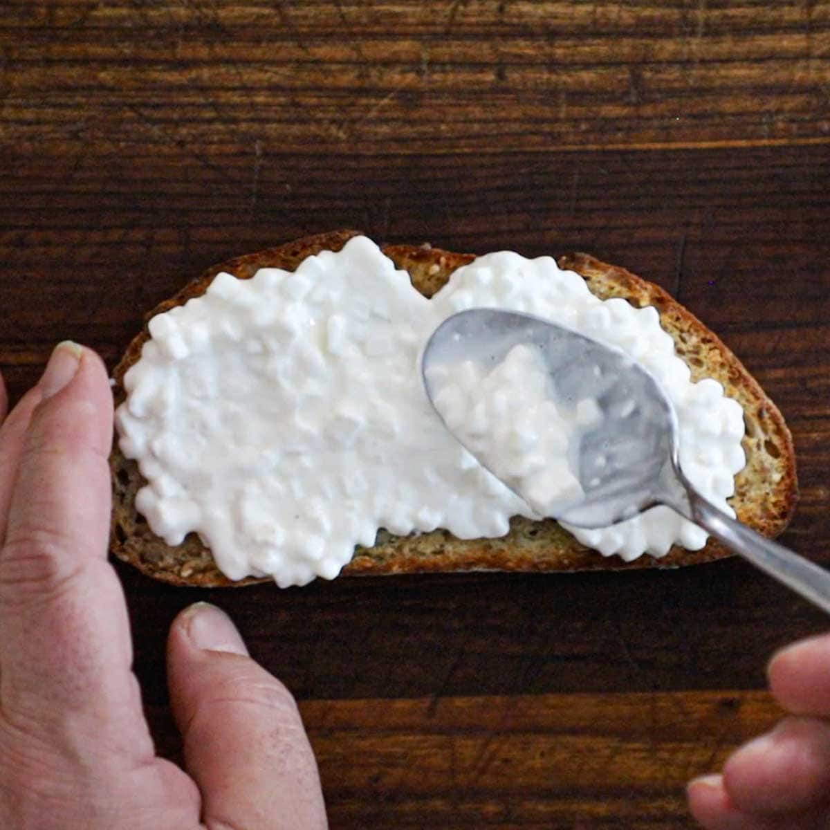 A person using a spoon to spread cottage cheese across the top of a toasted slice of multi grain bread on a wooden cutting board.