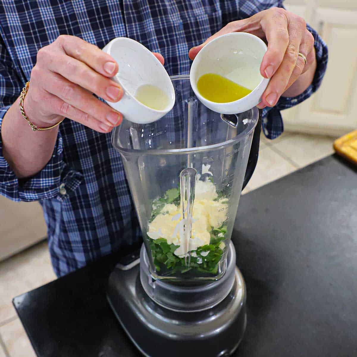 A person holding two small bowls of lemon juice and extra-virgin olive oil over a blender filled with cilantro, jalapeños, mayonnaise, sour cream, and garlic.