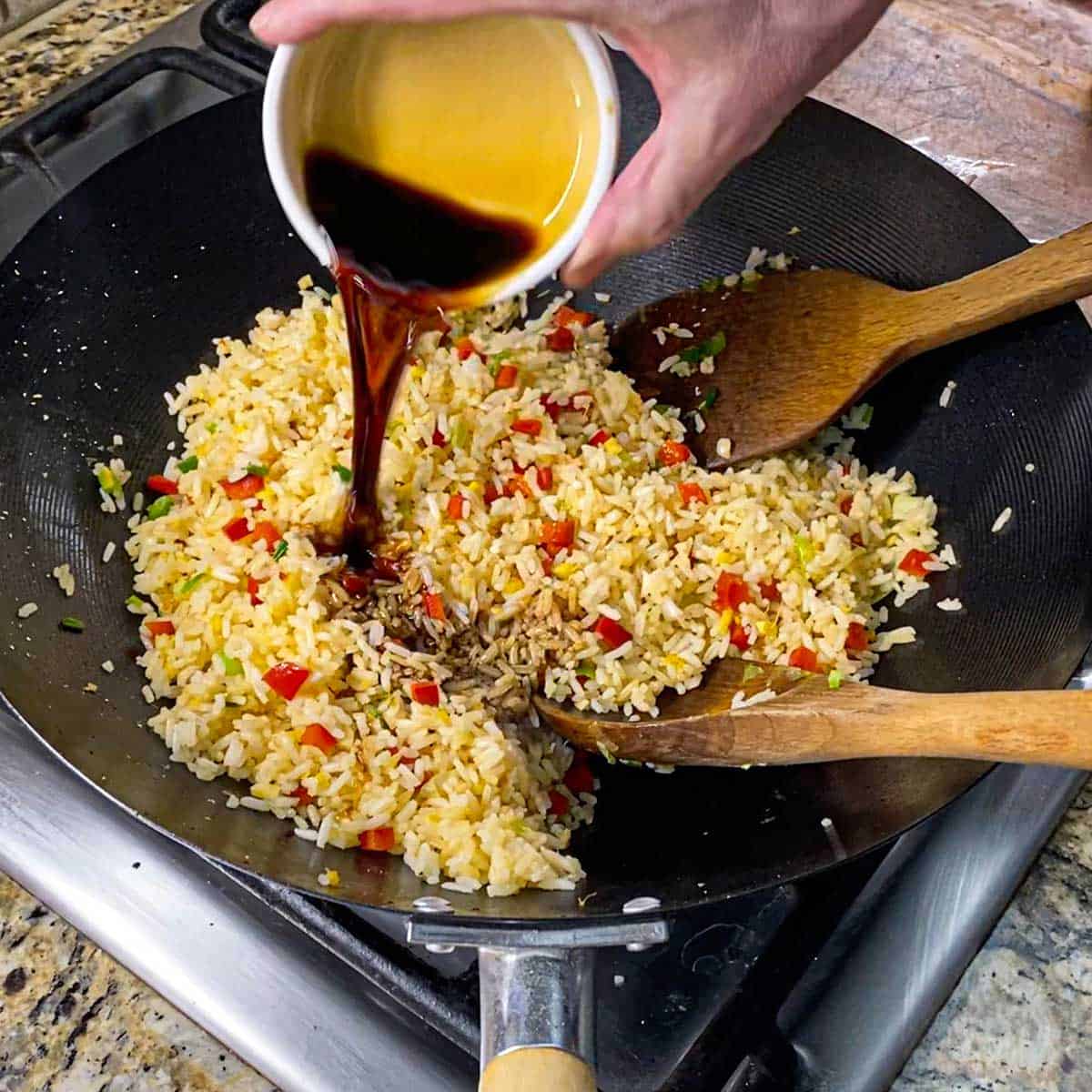 A person pouring soy sauce from a small white bowl into a wok that is filled with Peruvian fried rice that is being stir-fried with two large wooden spoons.