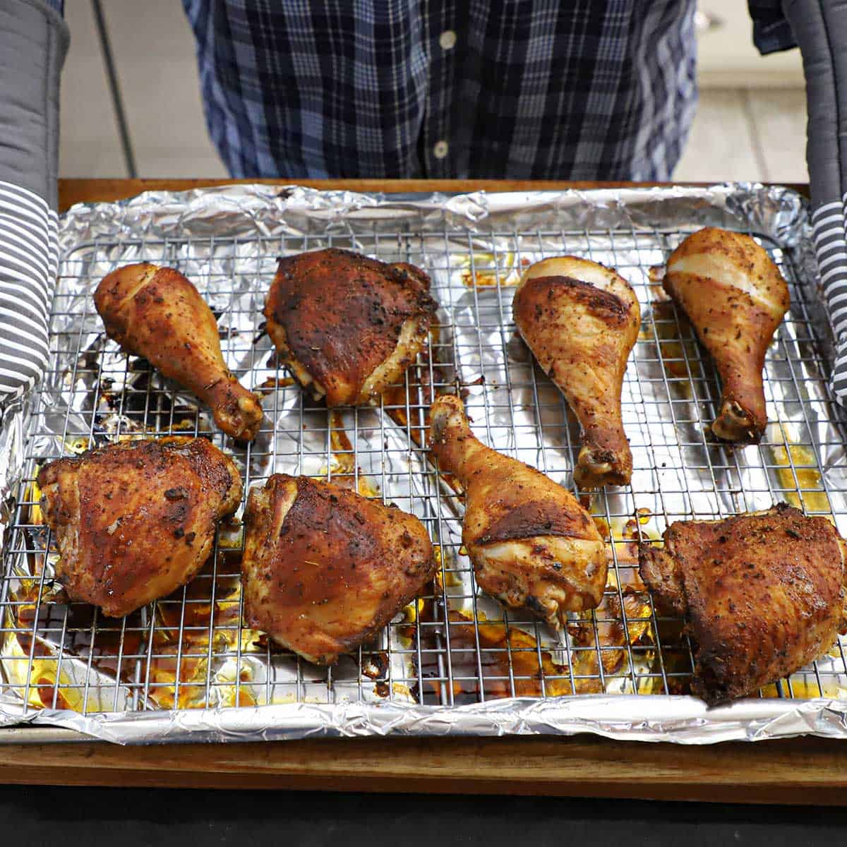 A person wearing oven mitts holding a baking sheet that is lined with foil and a baking rack holding freshly roasted Peruvian chicken pieces.
