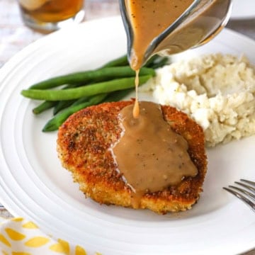 A person pouring brown gravy from a glass gravy boat over the top of a crispy pan-fried pork chop on a white dinner plate also containing a pile of mashed potatoes and steamed green beans.