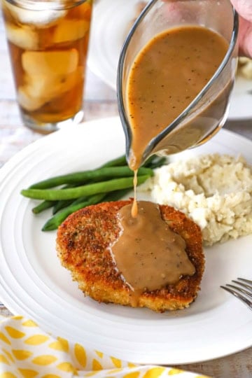 A person pouring brown gravy from a glass gravy boat over the top of a crispy pan-fried pork chop on a white dinner plate also containing a pile of mashed potatoes and steamed green beans.