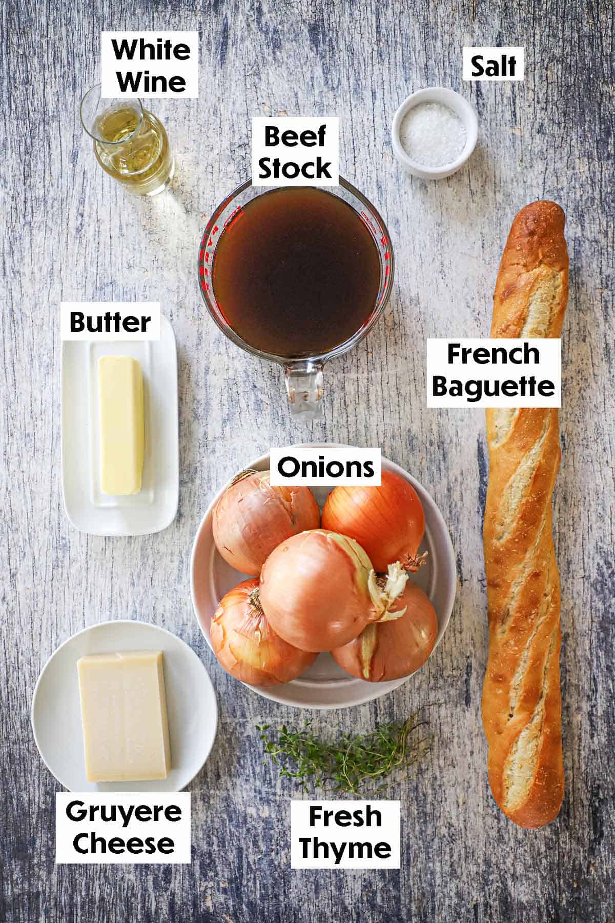 An arrangement of ingredients for homemade French onion soup on a grey wooden background including beef stock, onions, white wine, butter, Gruyere cheese, fresh thyme, a baguette, and salt.