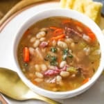 An overhead view of a white soup bowl filled with Classic Ham and Bean Soup (With Navy Beans) sitting on a white plate with a gold spoon resting nearby.