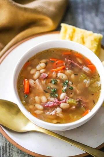 An overhead view of a white soup bowl filled with Classic Ham and Bean Soup (With Navy Beans) sitting on a white plate with a gold spoon resting nearby.