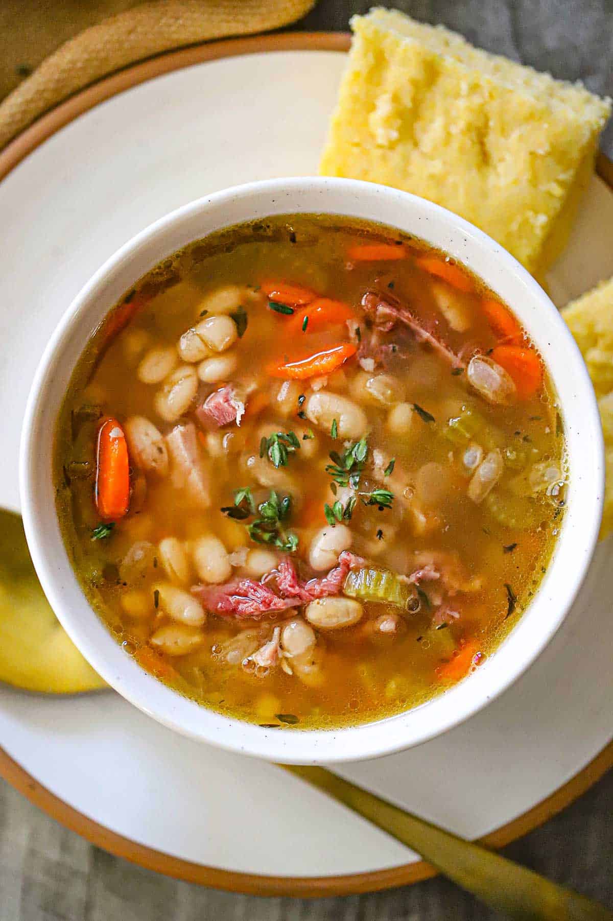 An overhead view of a white soup bowl filled with Classic Ham and Bean Soup (With Navy Beans) sitting on a white plate with a gold spoon resting nearby.