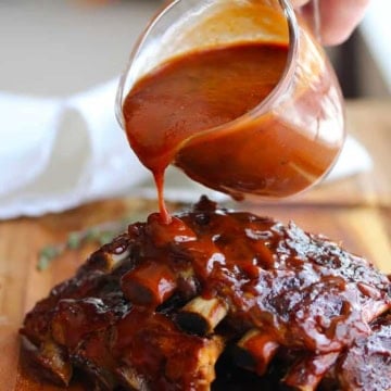 A person pouring homemade easy BBQ sauce over a pile of barbecued baby back ribs.