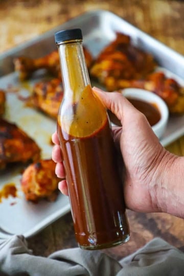 A person holding a tall thin bottle that is filled with easy BBQ sauce over a sheet pan filled with barbecue chicken.
