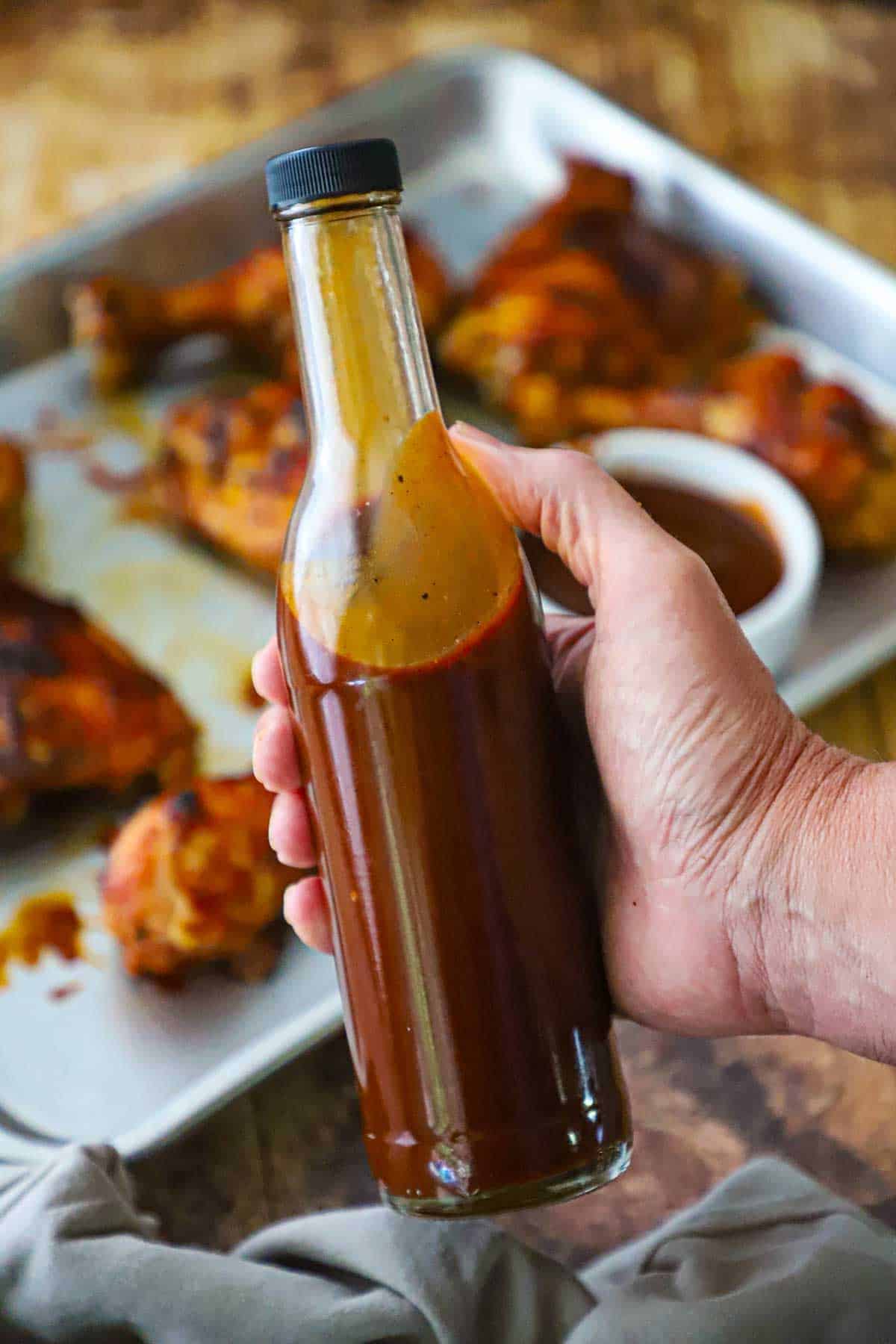 A person holding a tall thin bottle that is filled with easy BBQ sauce over a sheet pan filled with barbecue chicken.