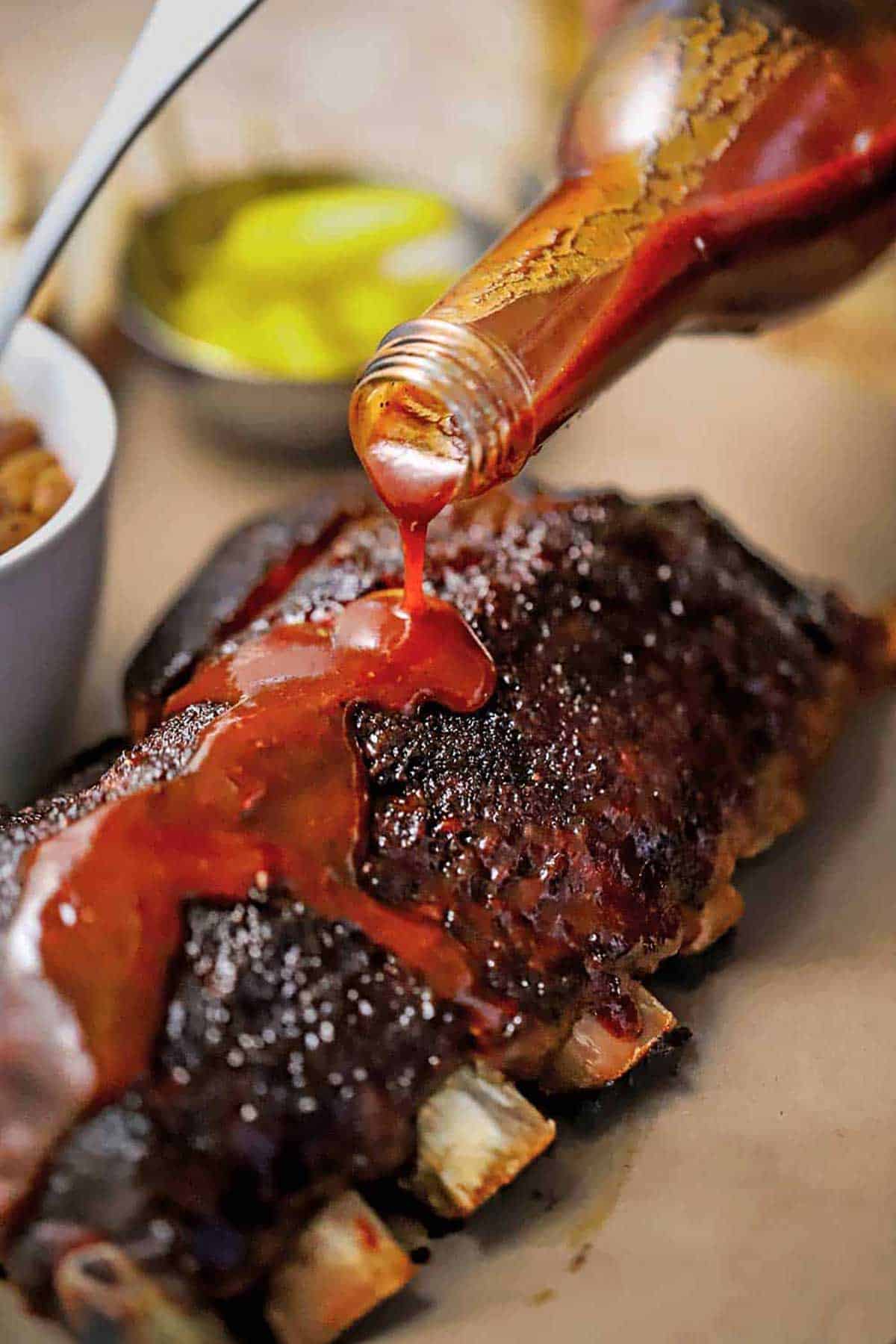 A person pouring homemade easy BBQ sauce over a rack of slow-cooker baby back ribs.