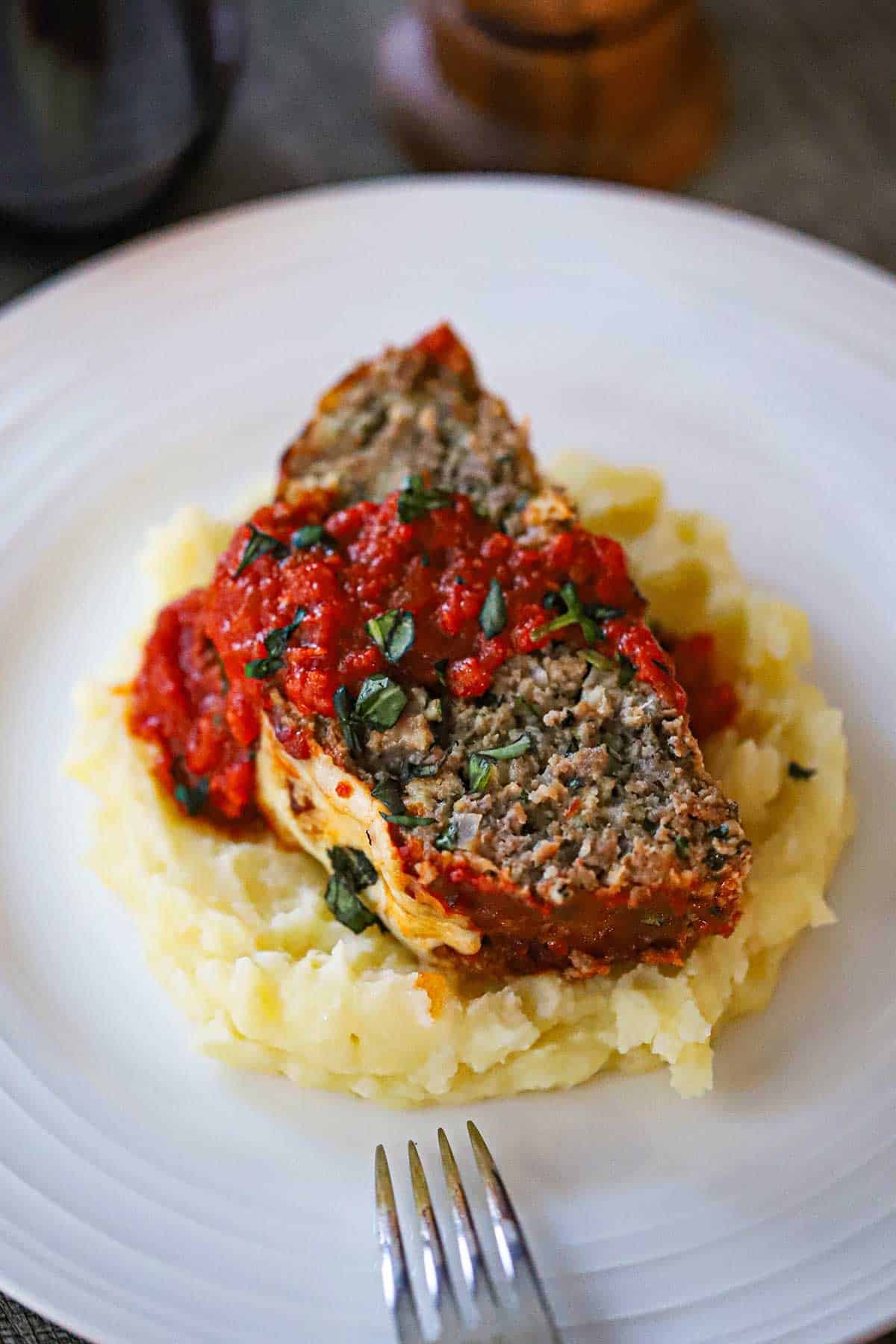 A slice of The Best Juicy Italian Meatloaf with Marinara Glaze resting on a pile of mashed potatoes on a white dinner plate.