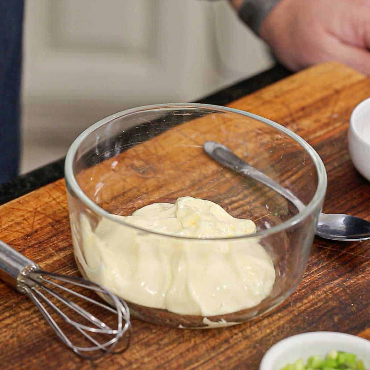 A person standing near a cutting board that has a glass bowl filled with a cup of mayonnaise in it.
