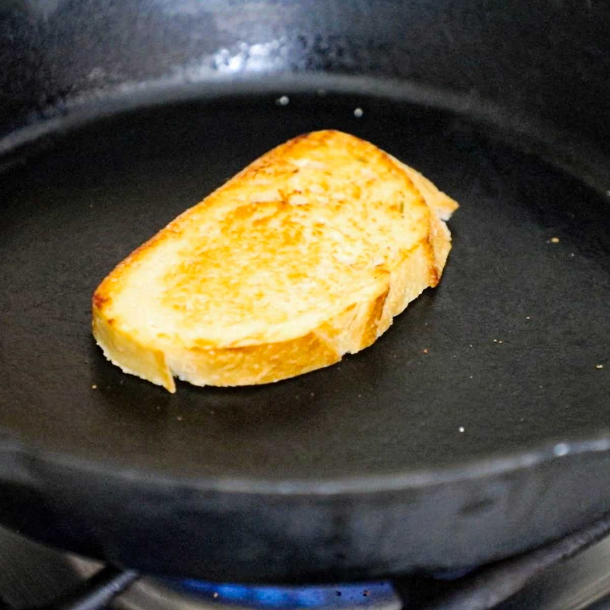 A slice of sourdough bread being toasted in a large cast-iron skillet over a gas stove.