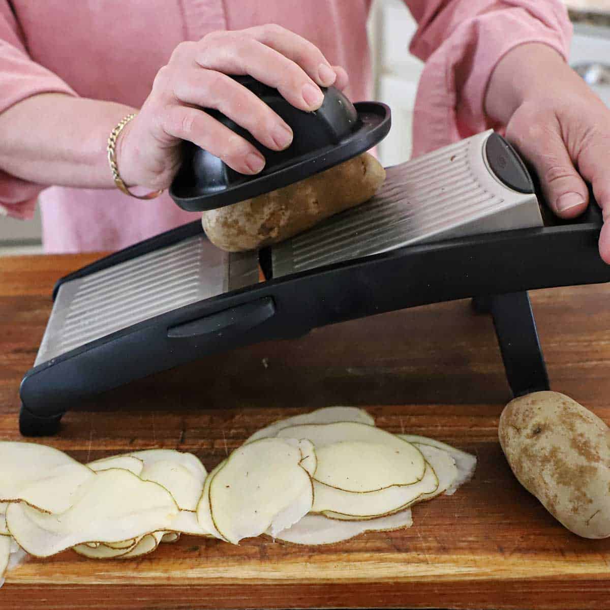 A person using a mandolin to slice a russet potato into paper thin slices on a wooden cutting board.