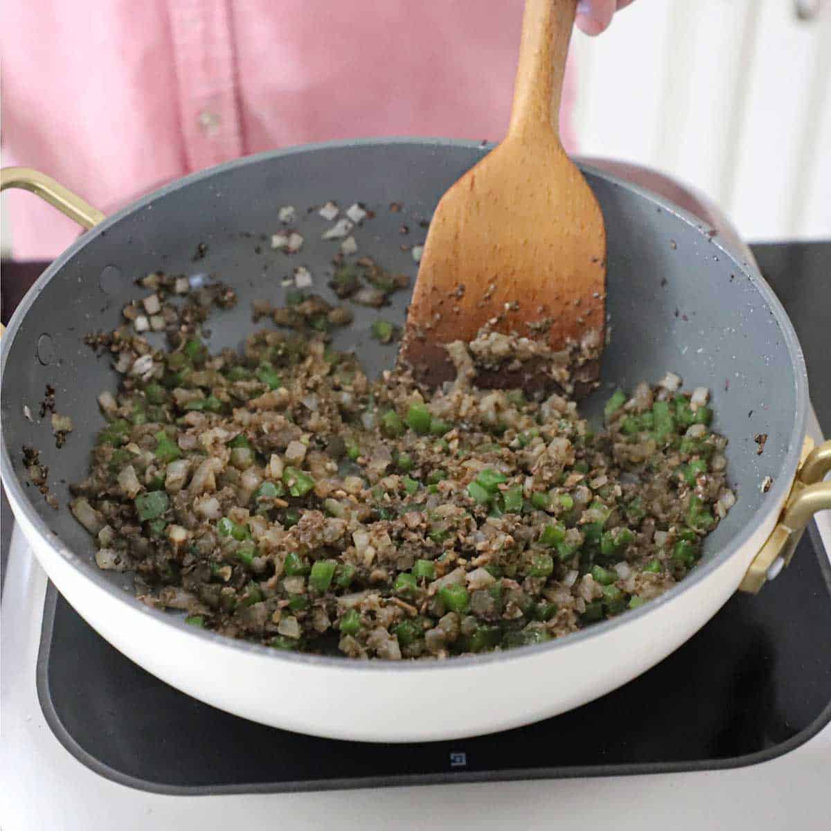A person using a large wooden spatula to stir sautéed onions with chopped green bell peppers and finely chopped mushroom in a large skillet.