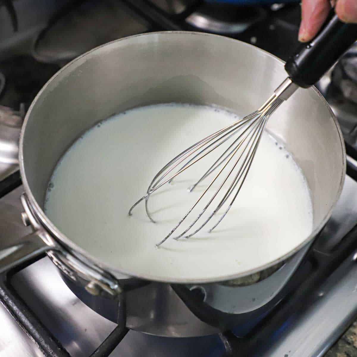 A person using a whisk to stir simmering milk on a gas stove.