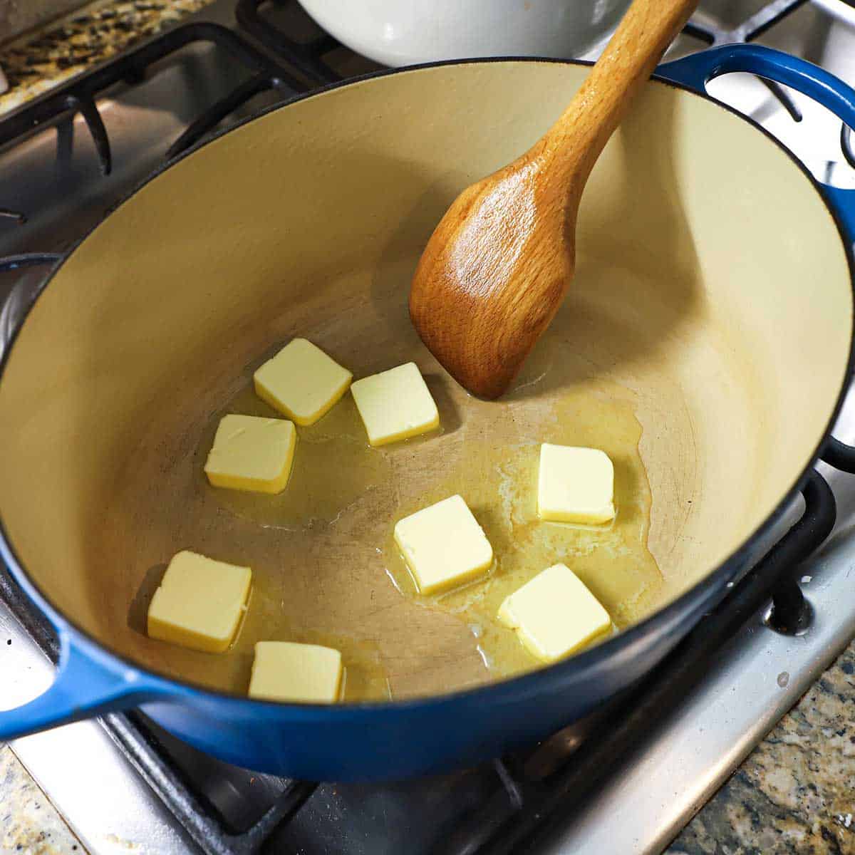 A person using a wooden spoon to stir pads of butter that are being melted in a large Dutch oven on a gas stove.