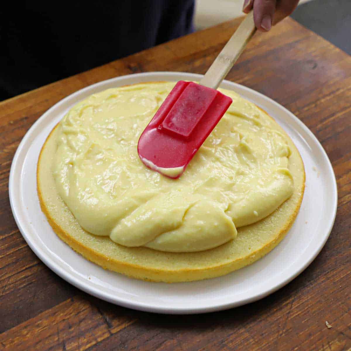 A person using a red rubber spatula to spread vanilla custard onto the surface of a thin sponge cake that is resting on a white circular plate on a cutting board.