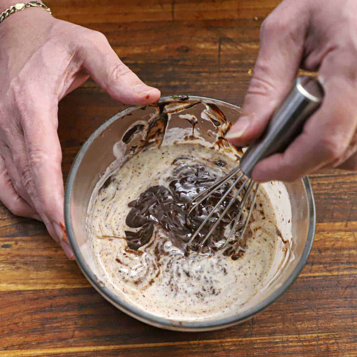 A person using a small whisk to combine hot cream with melting chunks of semi-sweet chocolate in a small glass bowl on a wooden cutting board.