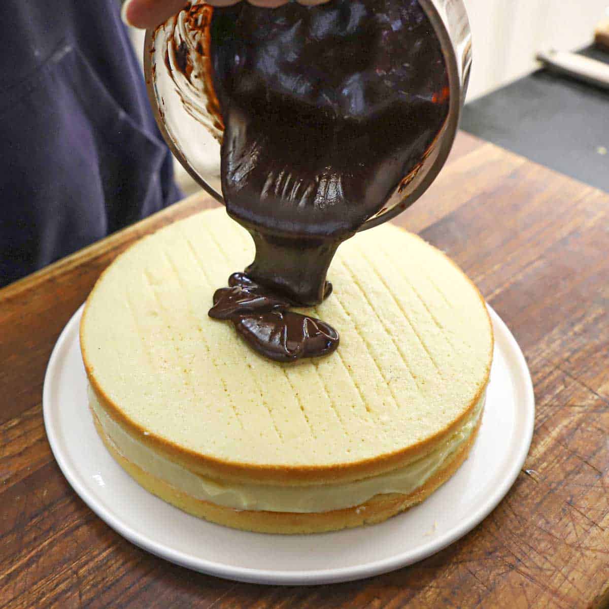 A person pouring a warm chocolate ganache mixture over the top of a freshly assembled Boston cream pie resting on a white plate on a wooden cutting board.
