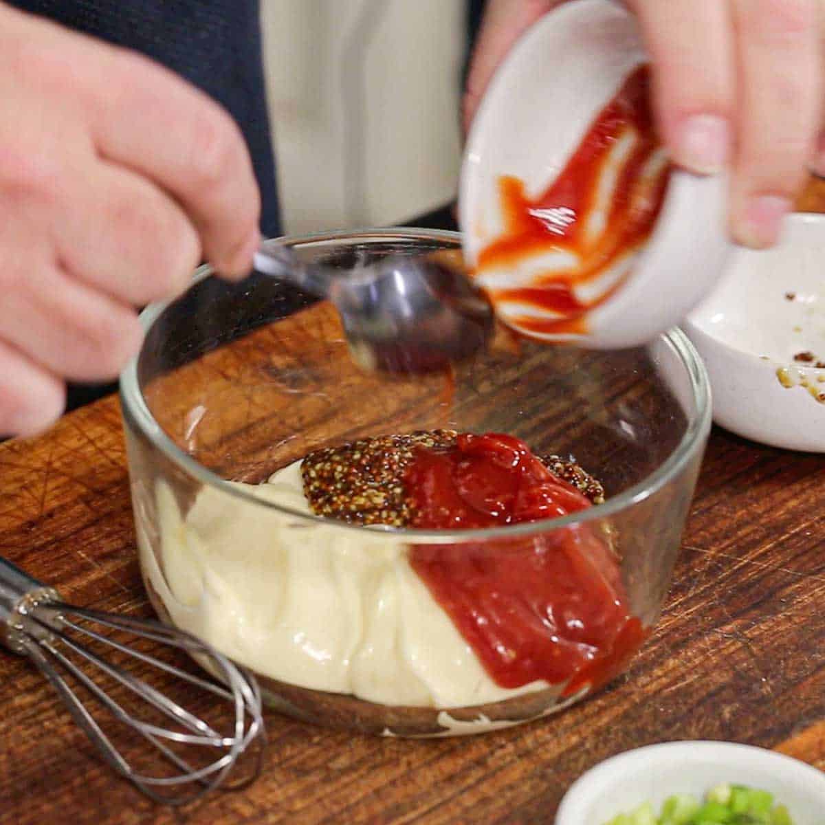 A person using a spoon to transfer ketchup from a small white bowl into a glass bowl that is filled mayonnaise and stone ground mustard.