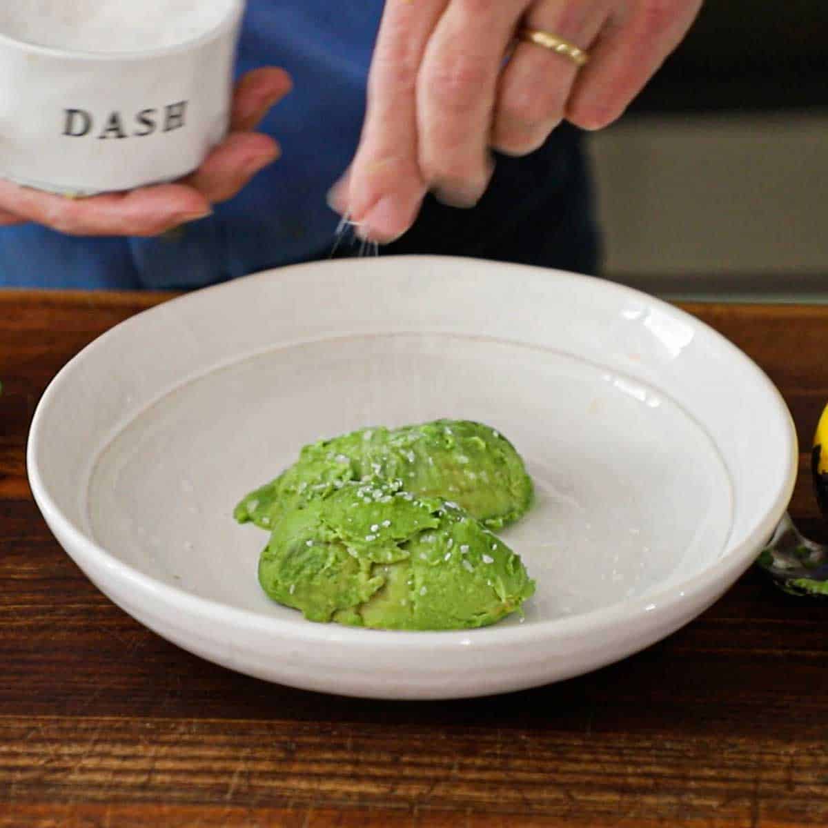A person sprinkling Kosher salt over two pieces of avocado flesh in a shallow white bowl.
