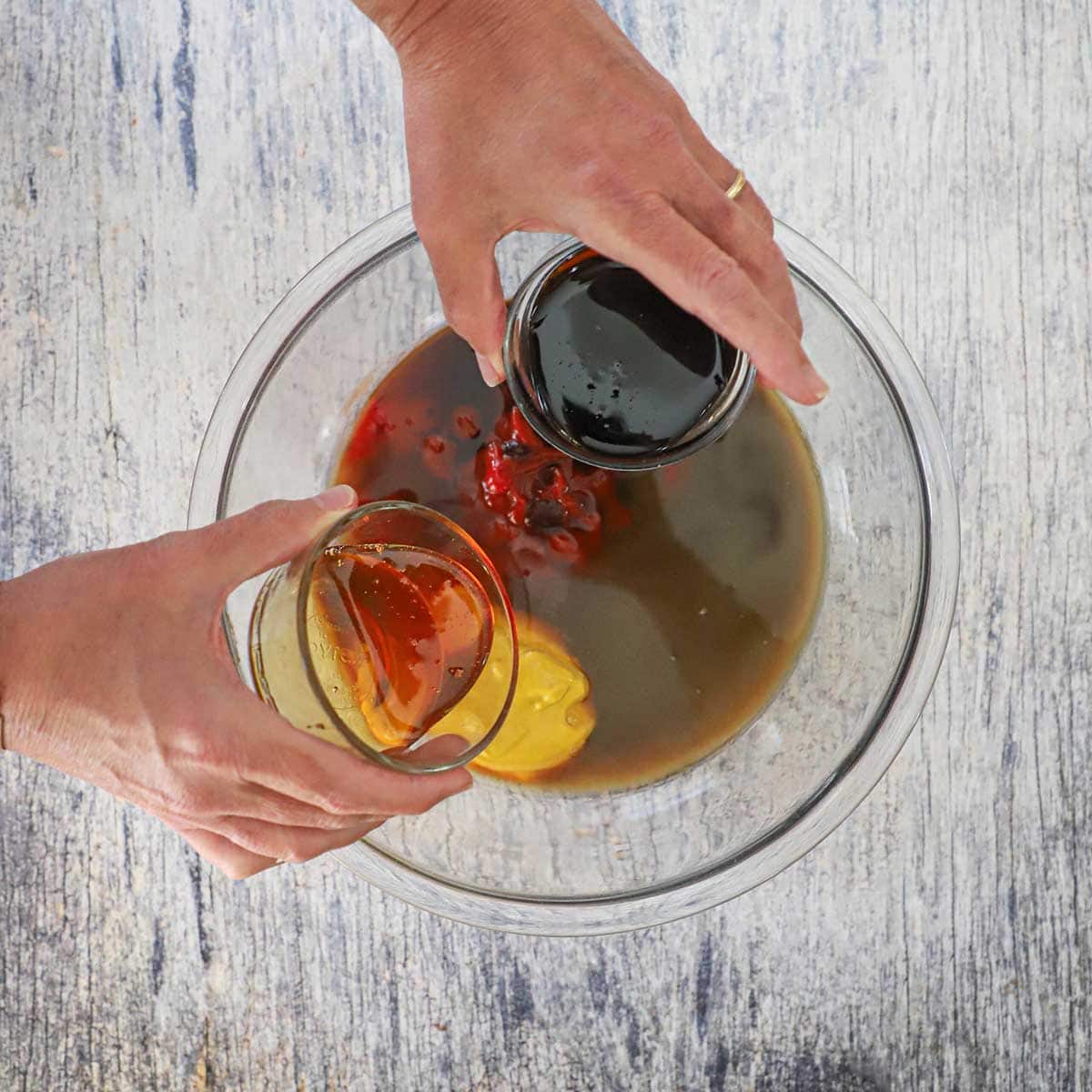 A person holding a small bowl of molasses in one hand and a small bowl filled with honey in the other hand both over a bowl filled with ingredients for easy homemade BBQ sauce.
