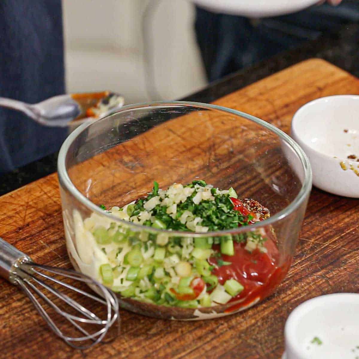 A glass bowl sitting on a wooden cutting board filled with portions of mayonnaise, ketchup, stone-ground mustard, minced garlic, chopped parsley, and chopped scallions.