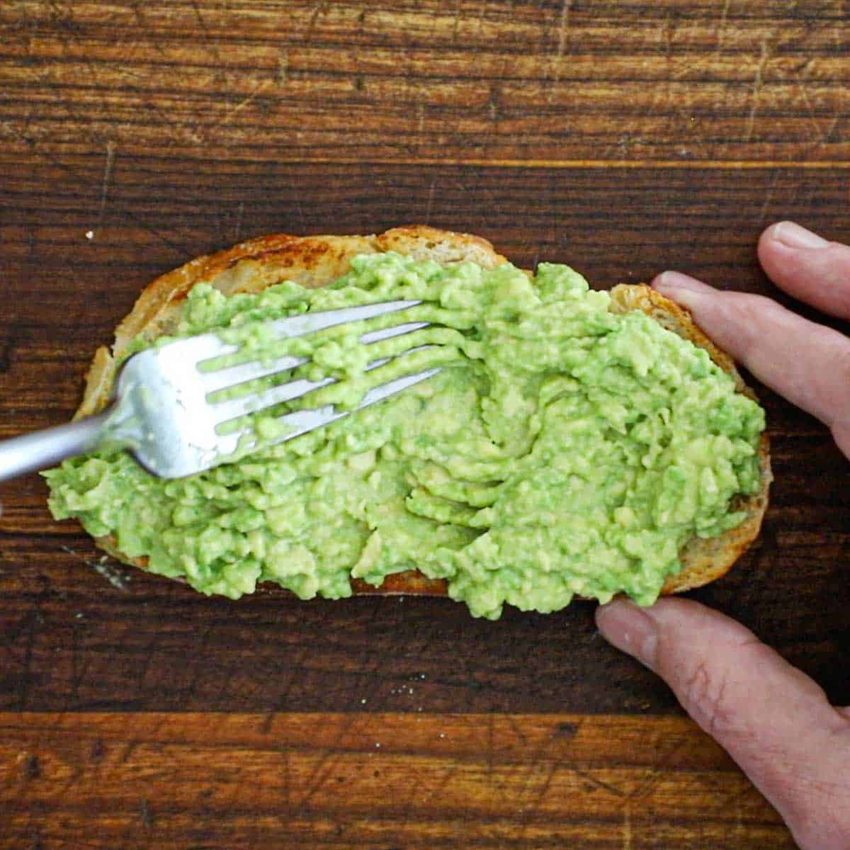 A person using a fork to spread smashed avocado flesh onto a sliced of toasted sourdough bread on a wooden cutting board.