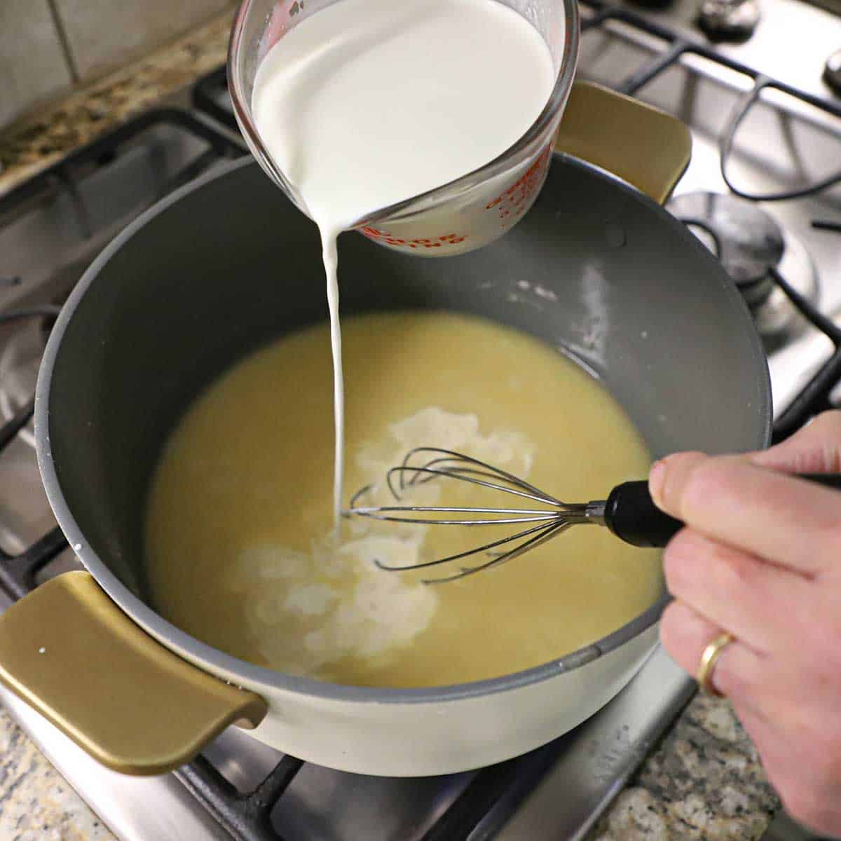A person using a whisk to stir chicken stock and half and half cream into a pot on a stove that is being thickened from a roux.