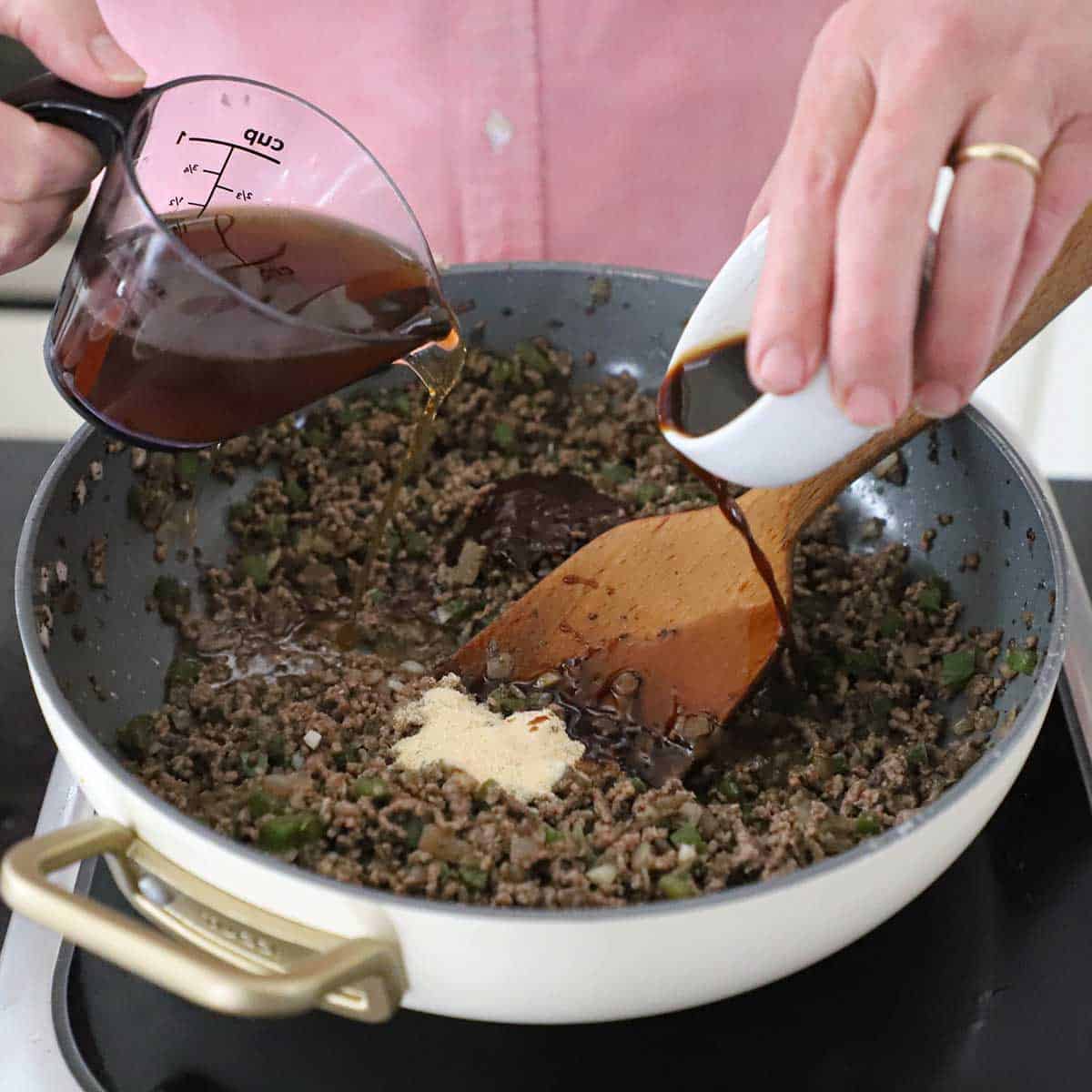 A person pouring steak sauce from a small white bowl in one hand and beef stock from a measuring up into a skillet filled with cooked ground beef with sautéed onions, green bell peppers, and onion powder.