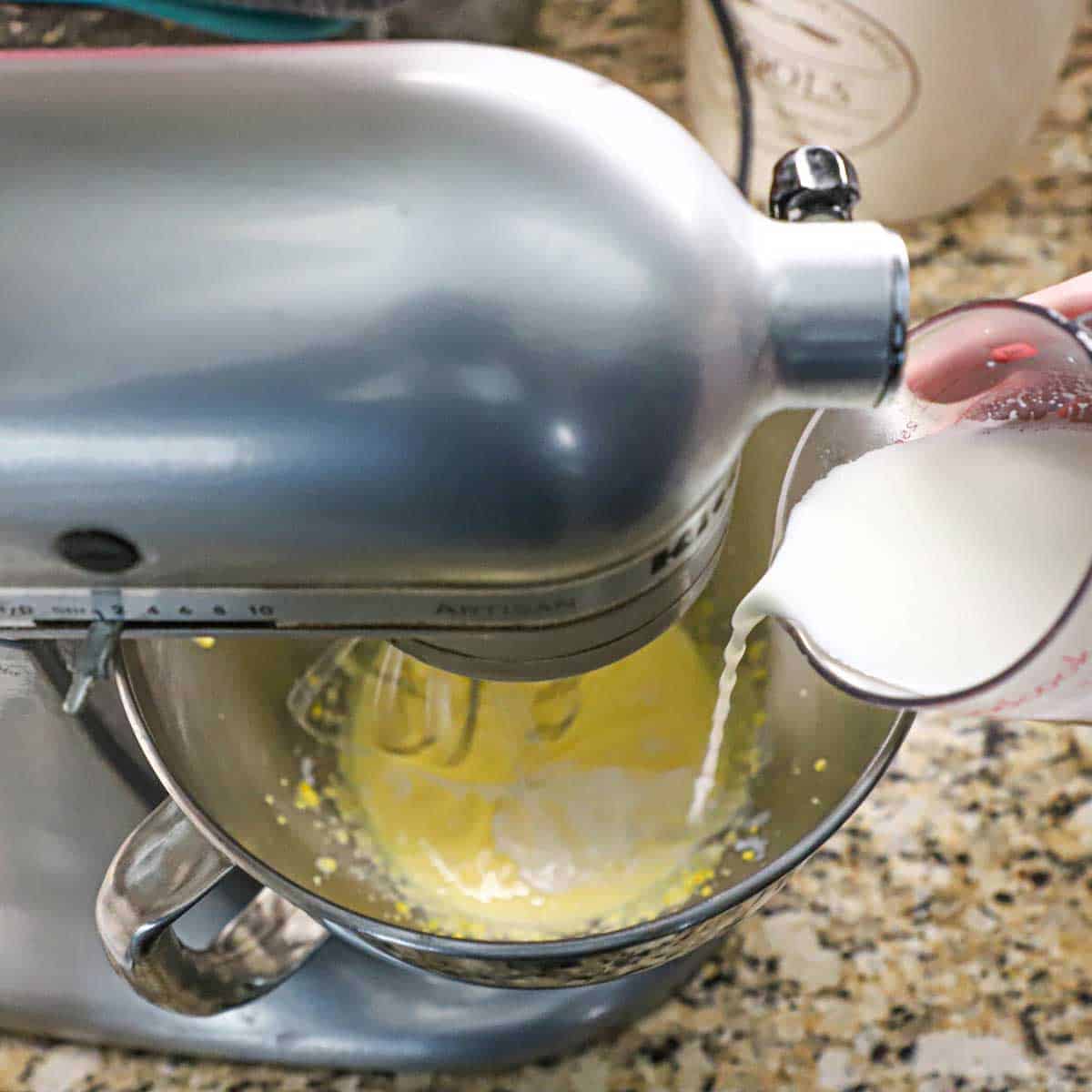 A person adding warm milk to creamed eggs that are being tempered in a stand-mixer.