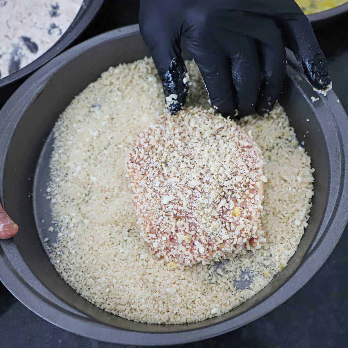 A person wearing a black latex glove dredging a boneless pork chop that a breadcrumb and Parmesan cheese mixture that is in a round baking pan.