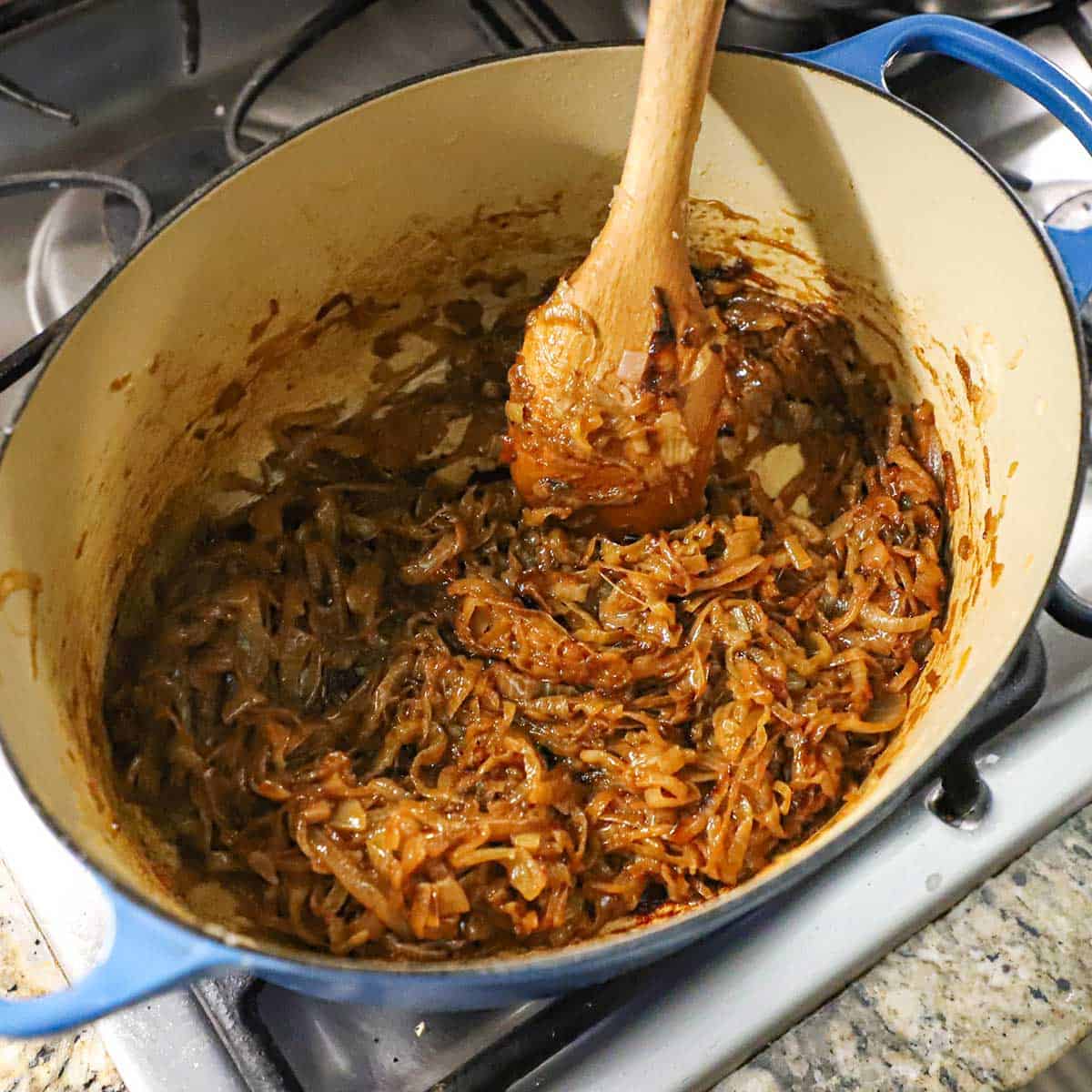 A large wooden spoon being used to stir deeply caramelized onions in a large Dutch oven on a gas stove.