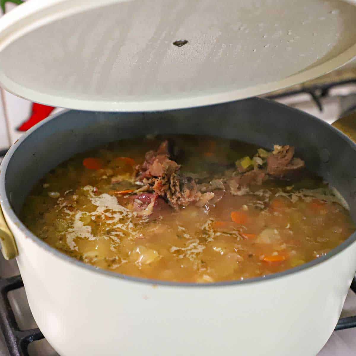 A person lifting the lid off of a large pot filled with simmering classic ham and beans soup with a ham bone visible in the middle of the pot.