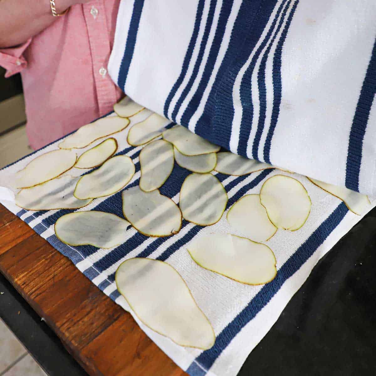 A person using a blue and white striped kitchen towel to dry thinly cut potato slices on a wooden cutting board.