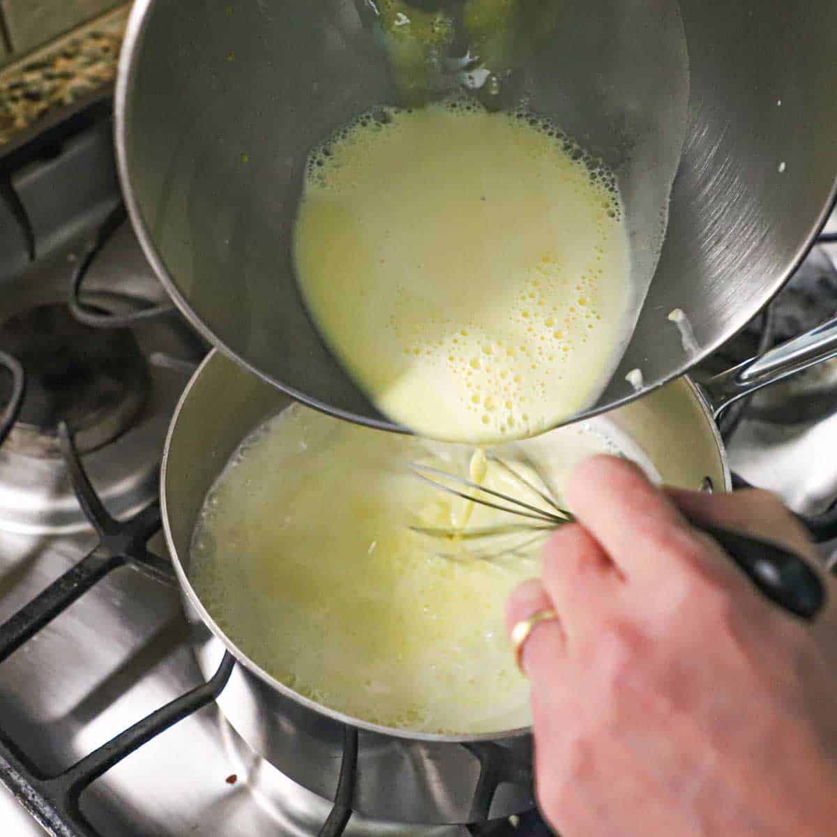 A person pouring tempered eggs from a large mixing bowl into a silver saucepan with simmering cream in it on a gas stove.