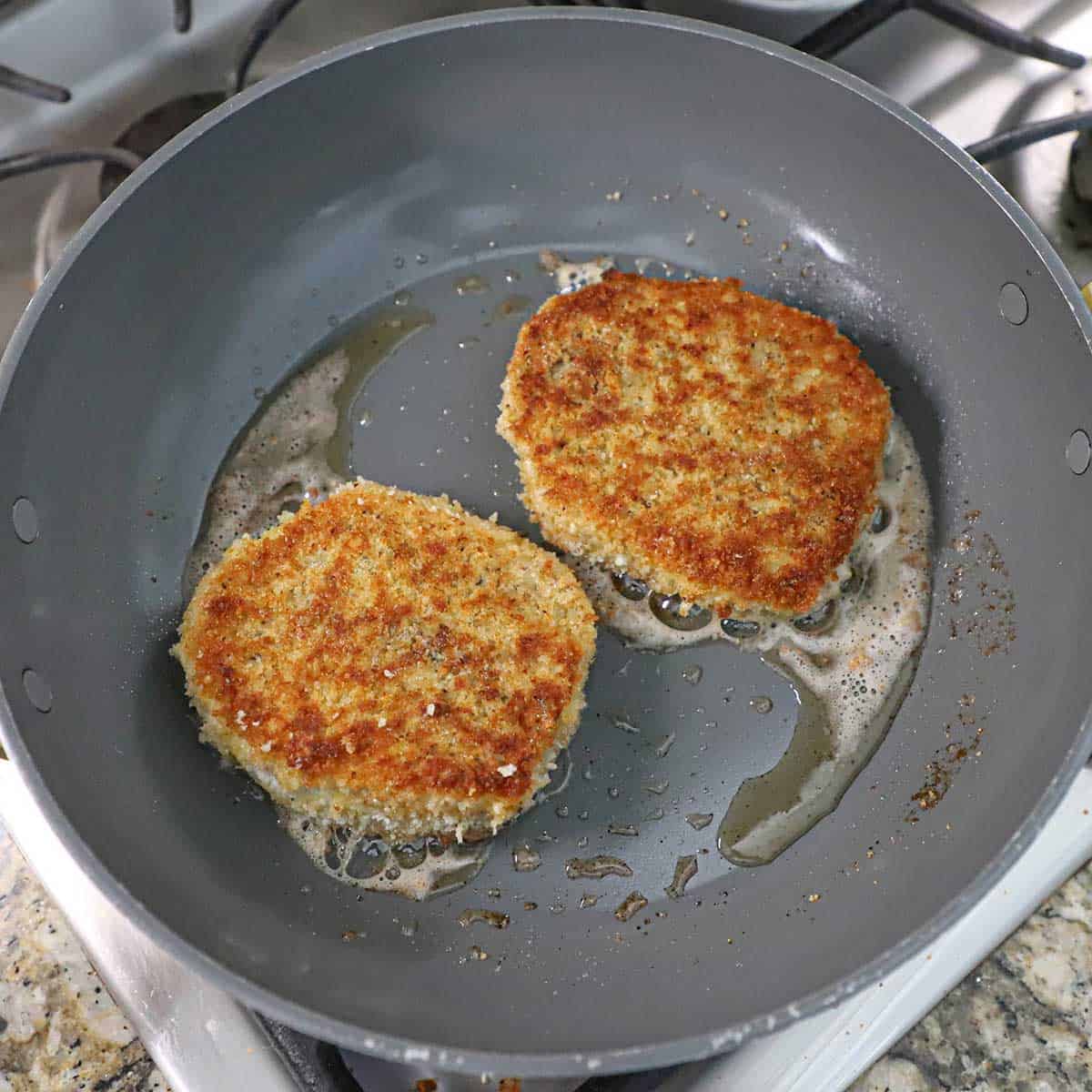 Two breaded boneless pork chops being pan-fried in a large non-stick skillet with a oil and butter on a gas stove.