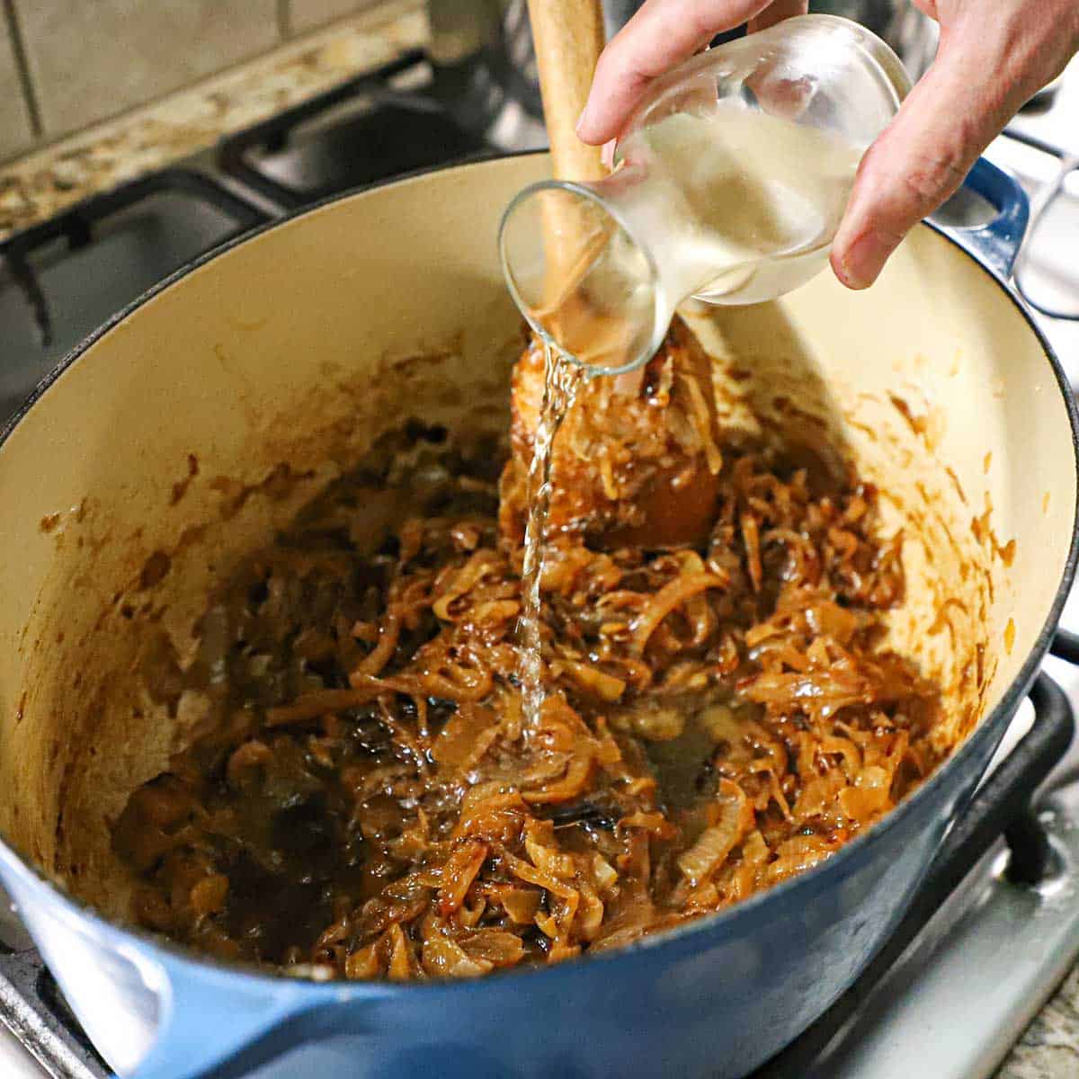 A person pouring white wine from a small glass carafe into a large Dutch oven that is filled with deeply caramelized onions simmering over a gas stove.