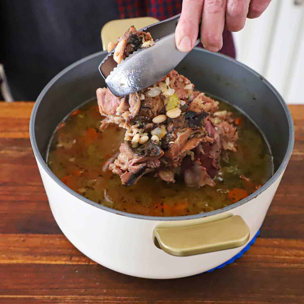 A person using a pair of metal tongs to lift a ham bone from a stock pot filled with classic ham and bean soup (with navy beans).